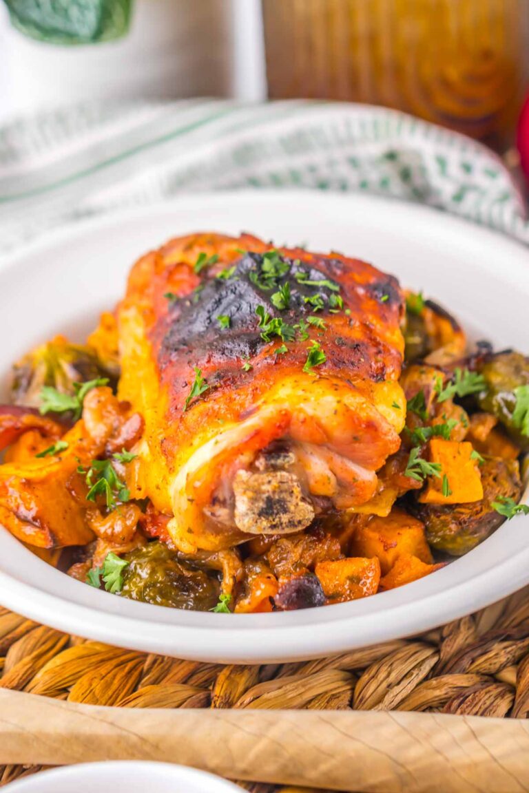 A roasted pork knuckle on a white plate, garnished with chopped parsley, sits atop a bed of roasted vegetables, including Brussels sprouts and sweet potatoes, reminiscent of the hearty goodness found in a Maple Dijon Chicken Sheet Pan dinner.