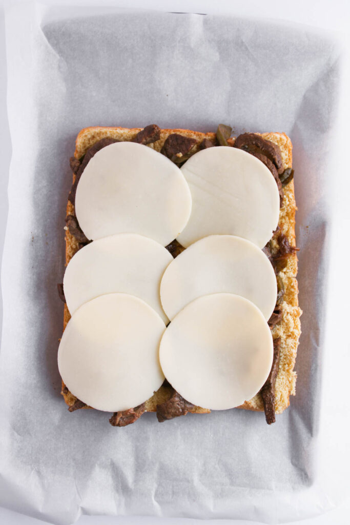 An open-faced sandwich on parchment paper topped with sliced cheese and saut&eacute;ed vegetables, reminiscent of Philly Cheesesteak Sliders.