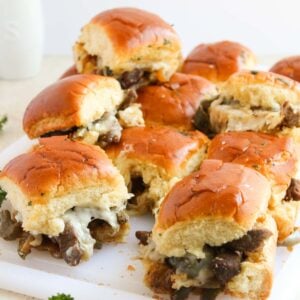 A pile of Philly Cheesesteak sliders filled with beef, cheese, and vegetables rests on a white cutting board, garnished with parsley.