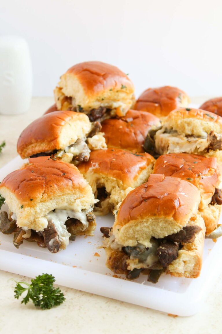 A pile of Philly Cheesesteak sliders filled with beef, cheese, and vegetables rests on a white cutting board, garnished with parsley.