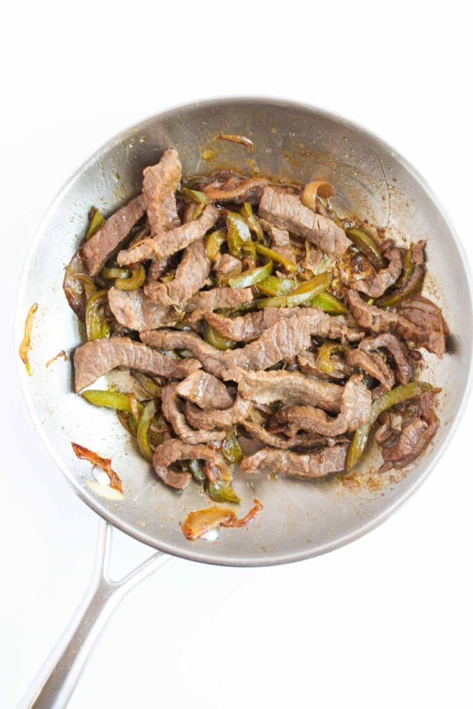 A skillet with saut&eacute;ed beef strips and green bell peppers evokes the tantalizing flavors of Philly Cheesesteak Sliders on a white background.
