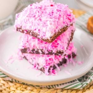 A stack of three pink Snoball Brownies, each coconut-covered square nestled on a white plate, rests elegantly on a woven mat.