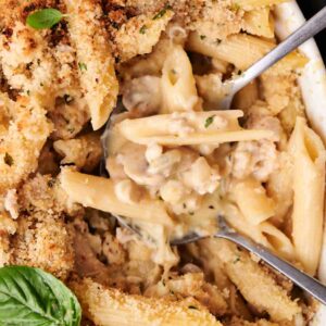 Close-up of baked pasta with creamy sauce, topped with breadcrumbs and herbs, garnished with fresh basil. A serving spoon is partially submerged in the dish.