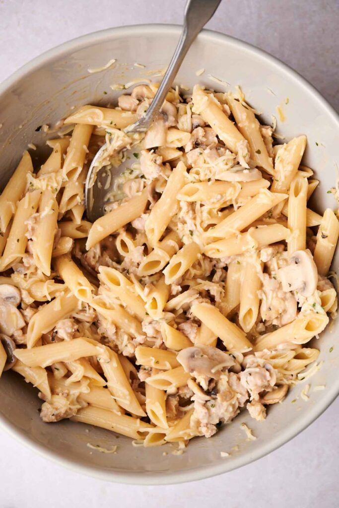 Bowl of penne pasta mixed with mushrooms and a creamy sauce, with a spoon.