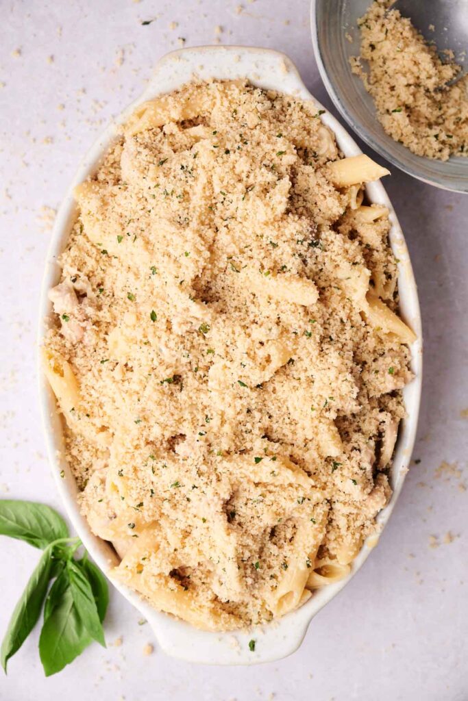 A casserole dish filled with penne pasta topped with a layer of seasoned breadcrumbs. A small bowl with breadcrumbs and a basil leaf are next to it.