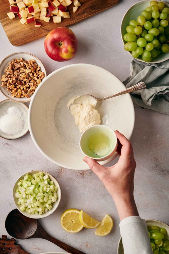 Hand holding a small bowl of liquid over a larger mixing bowl, surrounded by chopped apples, grapes, walnuts, celery, sugar, and lemon slices on a countertop.