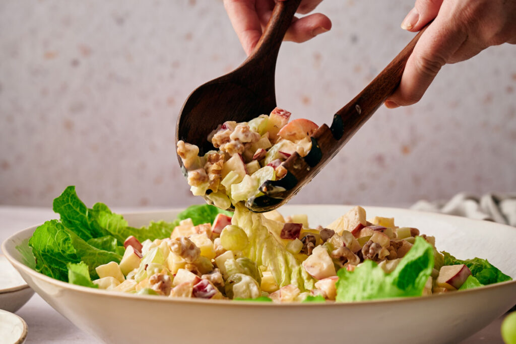 Close-up of a person serving salad with wooden utensils. The salad contains lettuce, apples, nuts, and other mixed ingredients in a white bowl.