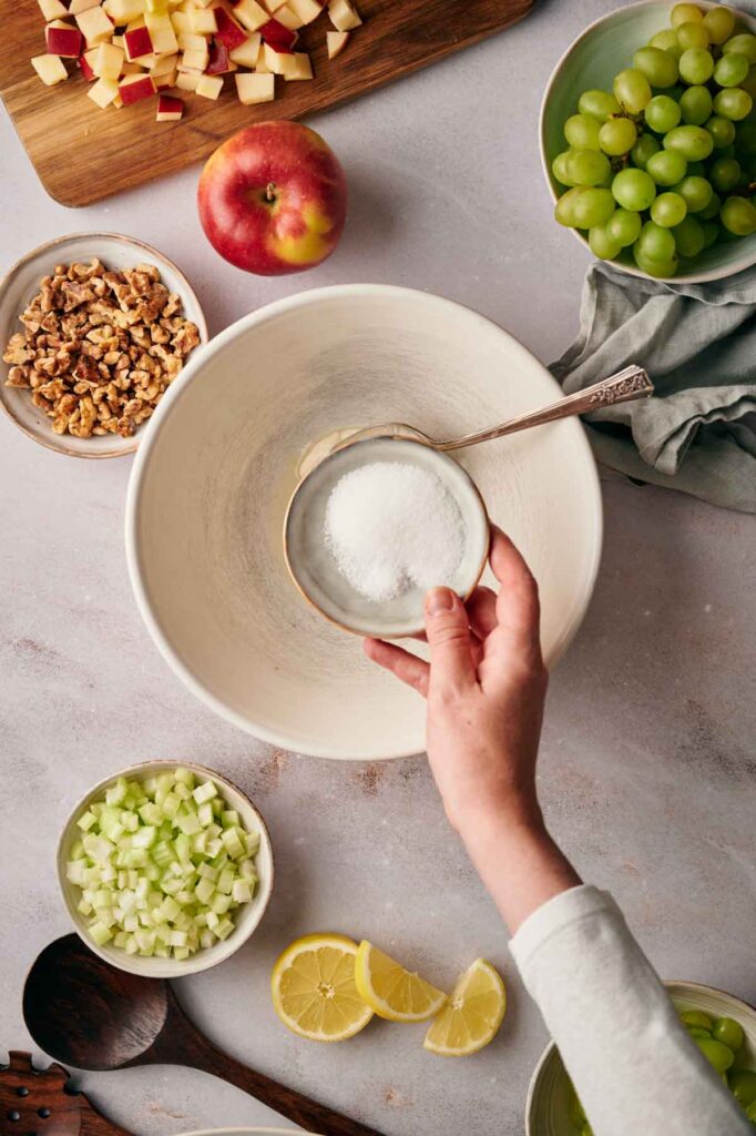 Hand holding a spoon of sugar above a mixing bowl surrounded by chopped apples, grapes, walnuts, celery, and lemon slices on a countertop.