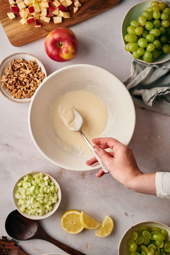 A person mixes batter in a bowl on a countertop surrounded by chopped apples, grapes, walnuts, celery, and lemon slices.
