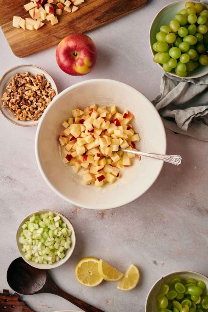 Ingredients for a Waldorf salad are arranged on a table: diced apples in a bowl, green grapes, walnuts, celery, a whole apple, sliced lemon, and a wooden spoon.