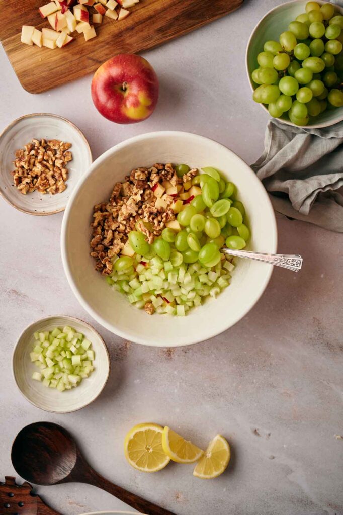 A bowl with chopped apples, walnuts, celery, and green grapes. Surrounding ingredients include sliced apples, walnuts, diced celery, a red apple, grapes, and lemon slices.