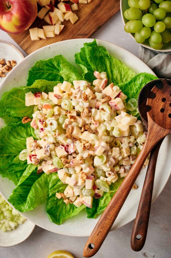 A Waldorf salad on a bed of lettuce, topped with diced apples, grapes, walnuts, and dressing. Wooden salad servers rest on the side. Apples and grapes are visible in the background.