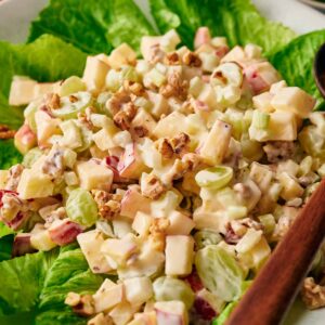A salad featuring chopped apples, grapes, walnuts, and diced cheese on a bed of lettuce, served in a bowl with a wooden spoon.