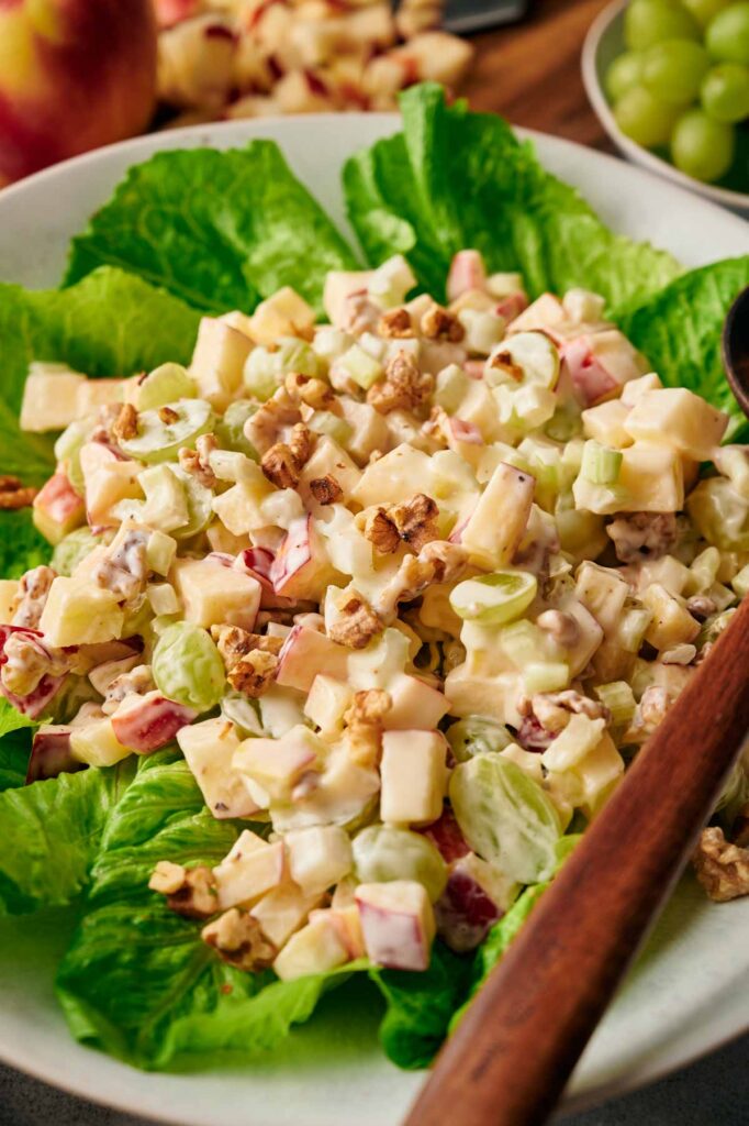 A salad featuring chopped apples, grapes, walnuts, and diced cheese on a bed of lettuce, served in a bowl with a wooden spoon.