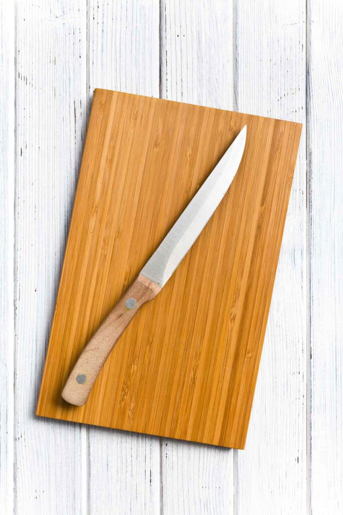 A knife with a wooden handle rests diagonally on a wooden cutting board, placed on a white wooden surface.