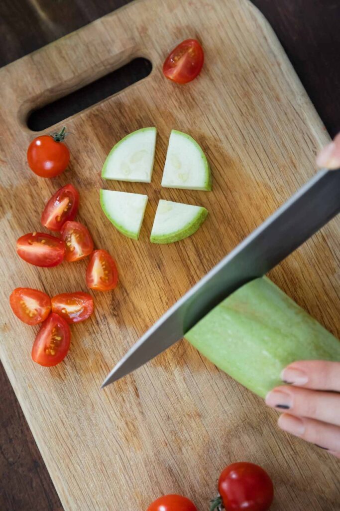A cutting board with sliced tomatoes and a green vegetable. A hand holds a knife, slicing a larger piece of the green vegetable.