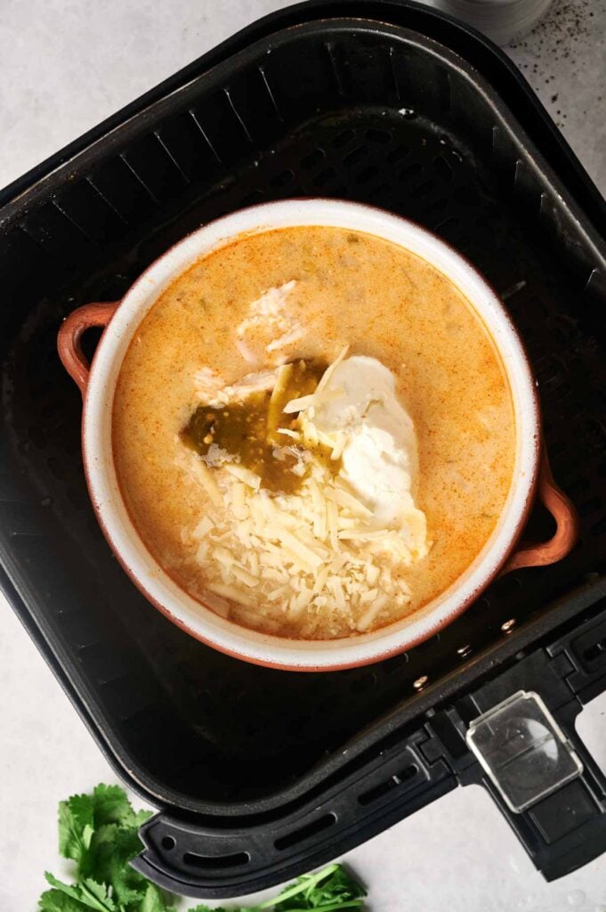 A bowl of creamy white chicken chili topped with melted cheese and sauce rests inside an air fryer basket.