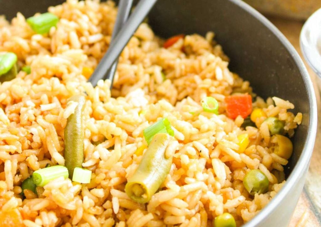 A close-up of a bowl of oven baked fried rice with green beans, peas, diced carrots, and chopped green onions, served with a black serving spoon.