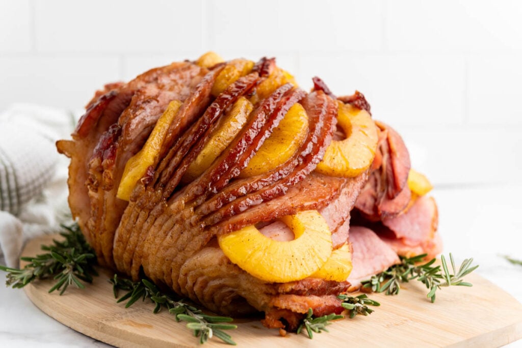 A brown sugar pineapple ham, spiral-sliced and garnished with pineapple rings and sprigs of rosemary, sits on a wooden cutting board.