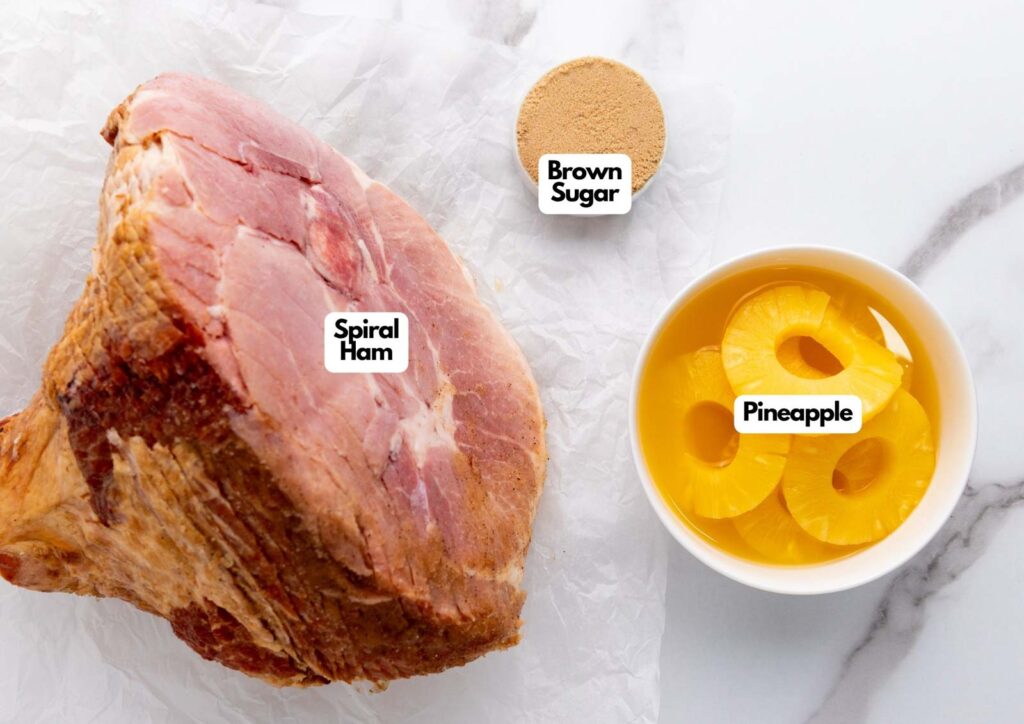 A spiral ham on parchment, a bowl of pineapple rings in juice, and a portion of brown sugar are displayed, perfect for making classic brown sugar pineapple ham. Each ingredient is clearly labeled.