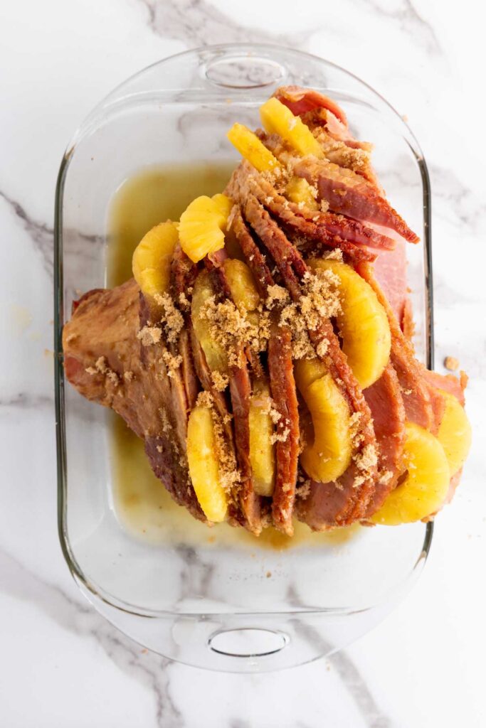In a glass baking dish, brown sugar pineapple ham is artfully arranged, with glazed ham slices topped by sweet pineapple rings and a sprinkle of brown sugar, all set on a sleek marble countertop.