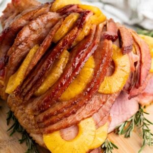Sliced ham topped with brown sugar-glazed pineapple rings and garnished with rosemary on a wooden board.