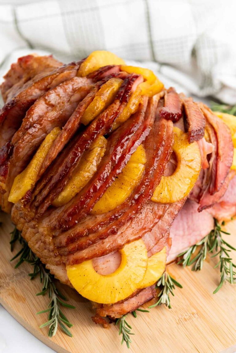 Sliced ham topped with brown sugar-glazed pineapple rings and garnished with rosemary on a wooden board.