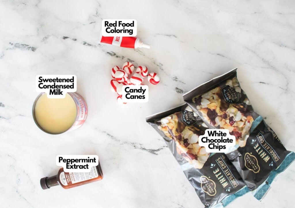 A can of sweetened condensed milk, peppermint extract, red food coloring, candy canes, and two bags of white chocolate chips are arranged on a white surface&mdash;everything you need to create festive candy cane fudge.