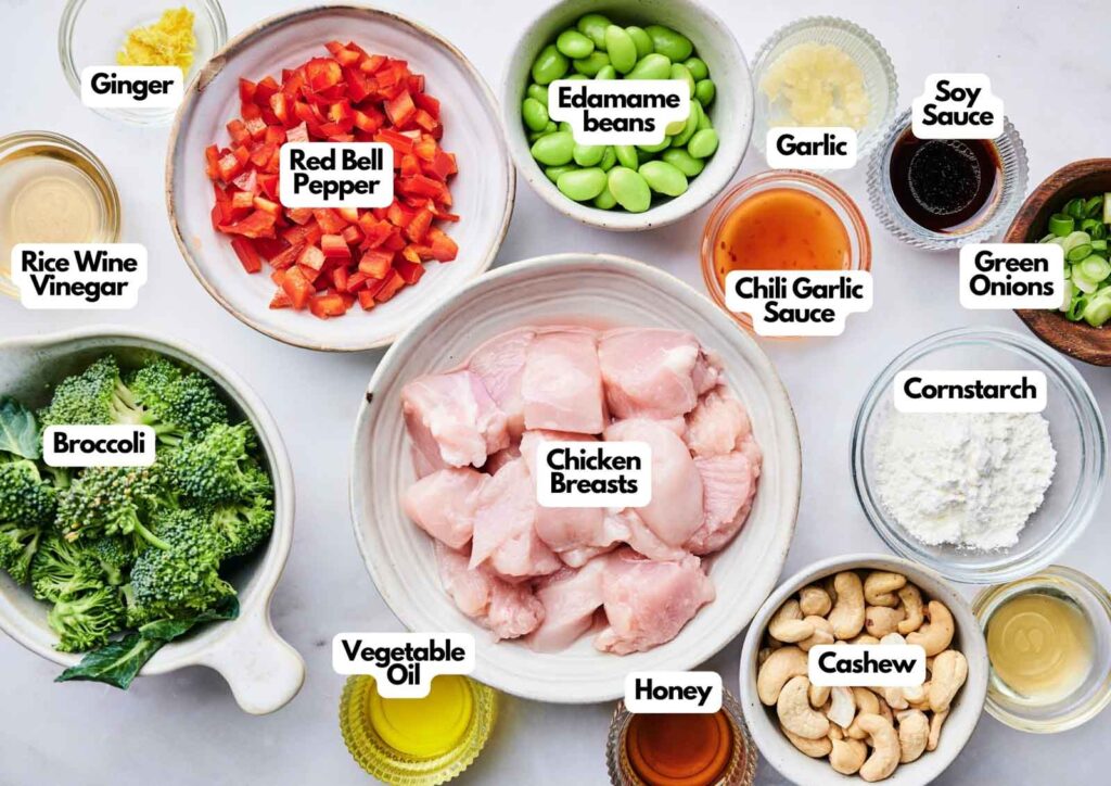 Ingredients for Cashew Chicken stir-fry&mdash;chicken breasts, red bell pepper, broccoli, edamame, cashews, sauces, cornstarch, and seasonings&mdash;are arranged in bowls on a white surface.