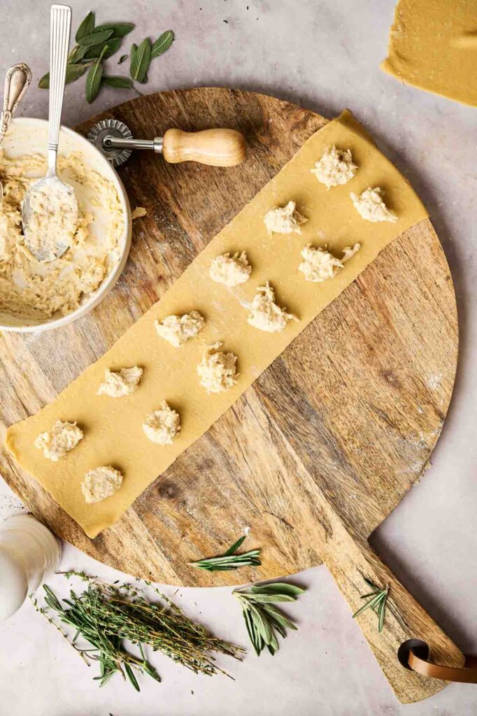 Wooden board with a strip of pasta dough topped with dollops of filling, surrounded by fresh herbs and a bowl containing the remaining mixture&mdash;perfectly illustrating how to make ravioli.