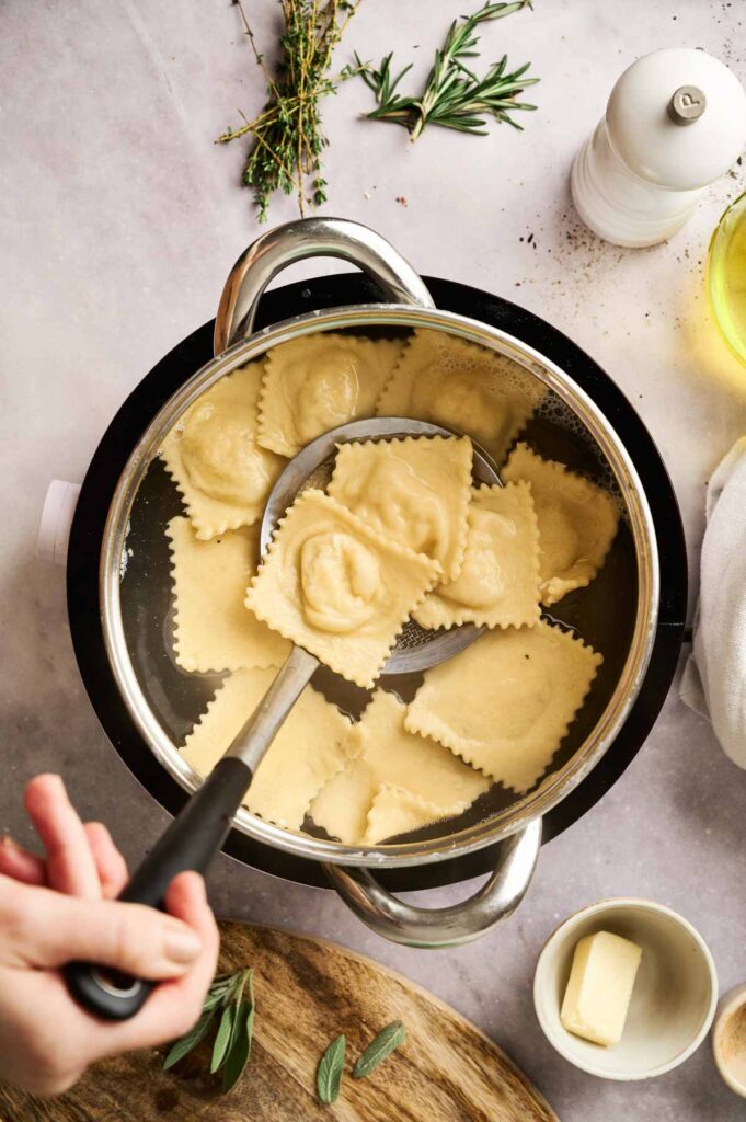 A pot of boiling water with ravioli being stirred by a slotted spoon, offering a glimpse of how to make ravioli just right. A small dish of butter and herbs sits nearby on the countertop, ready to enhance the flavors.