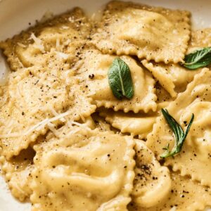 A plate of homemade ravioli topped with grated cheese, black pepper, and fresh sage leaves showcases how to make ravioli deliciously inviting.