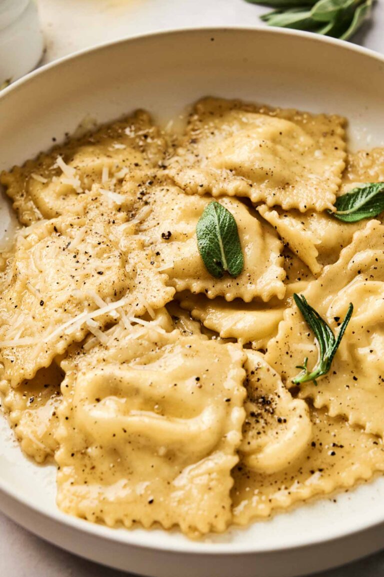 A plate of homemade ravioli topped with grated cheese, black pepper, and fresh sage leaves showcases how to make ravioli deliciously inviting.
