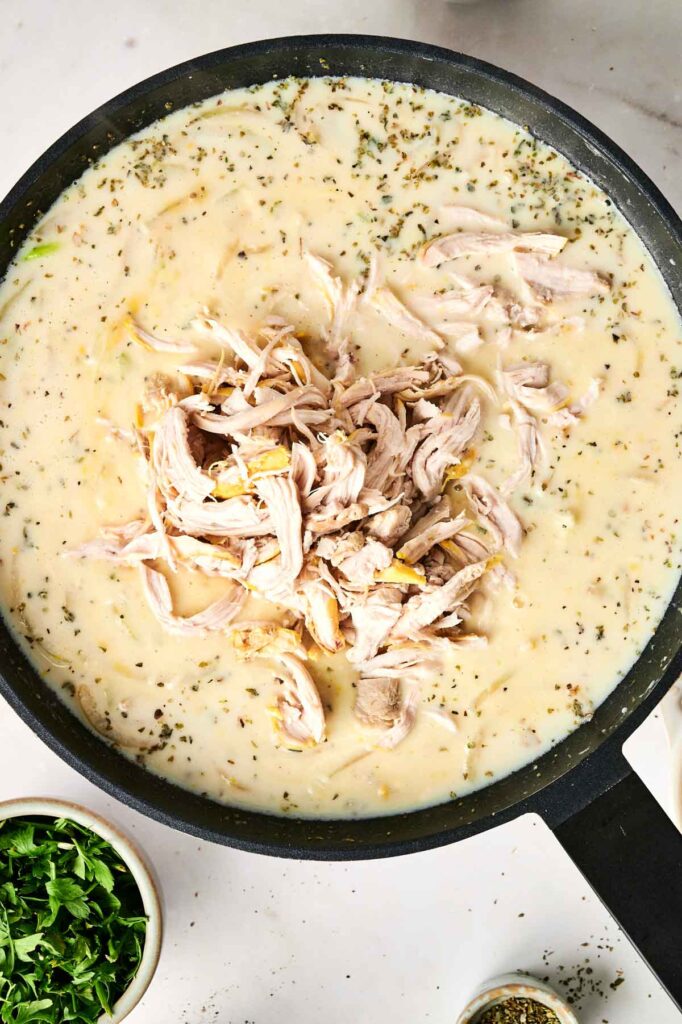 A pan filled with creamy chicken noodle casserole featuring shredded chicken and herbs. Small bowls with chopped herbs are visible nearby.