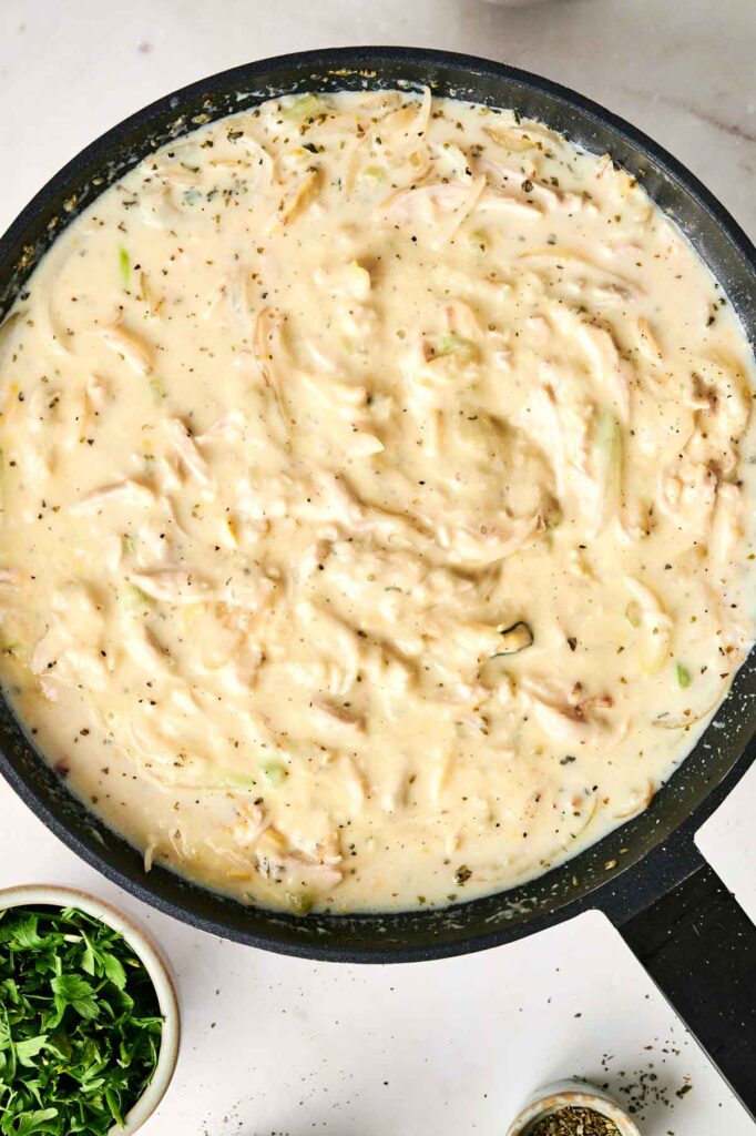 A skillet filled with creamy chicken noodle casserole and vegetable soup is surrounded by small bowls of chopped herbs and spices.