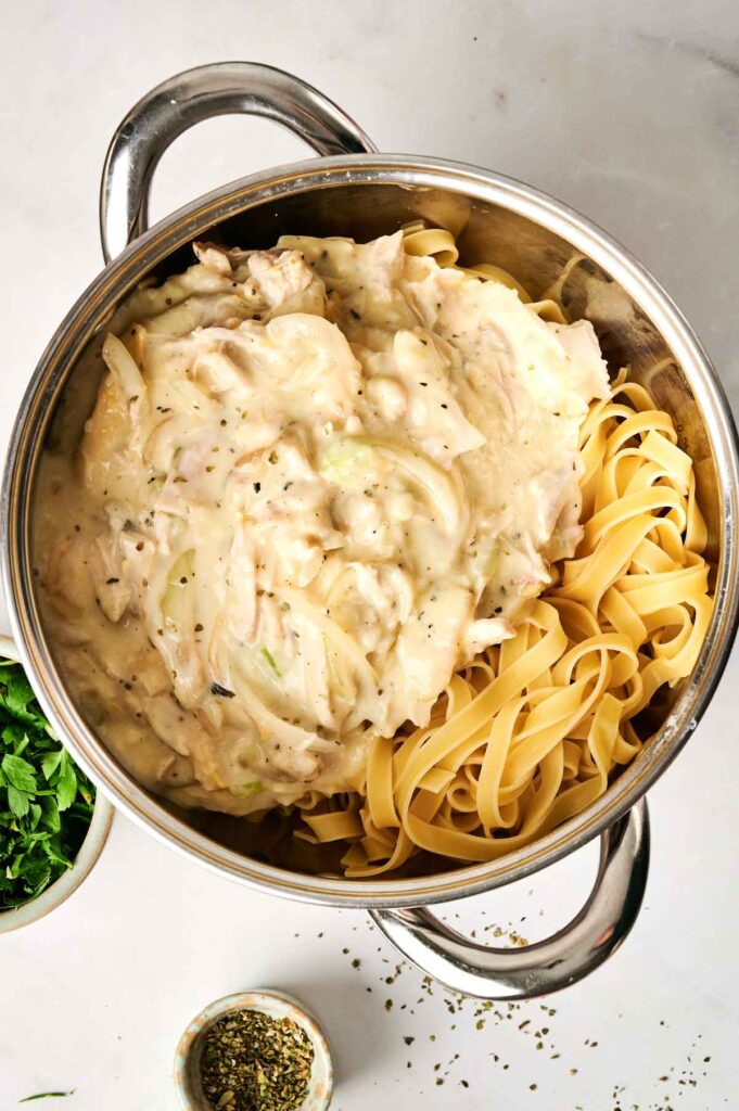A pot containing a hearty chicken noodle casserole with a creamy mixture, sprinkled with herbs. Parsley and seasoning sit in bowls nearby.