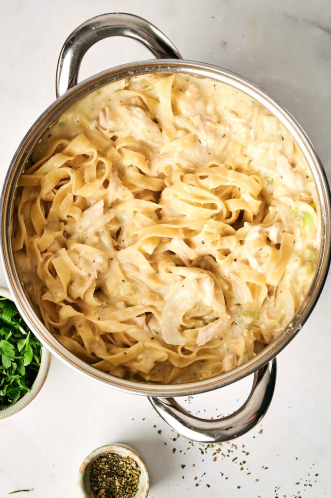 A pot of fettuccine pasta mixed with creamy sauce and reminiscent of a chicken noodle casserole, surrounded by small bowls of herbs on a white surface.