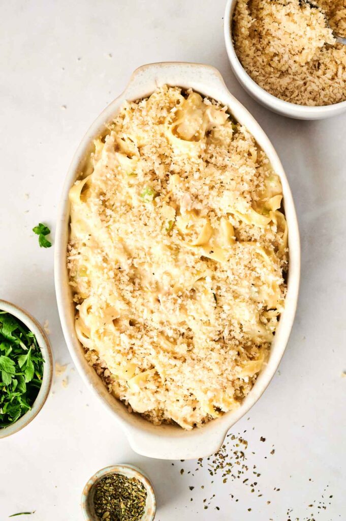 A delicious chicken noodle casserole dish filled with pasta and topped with breadcrumbs, surrounded by bowls of extra breadcrumbs, chopped herbs, and seasonings on a white surface.