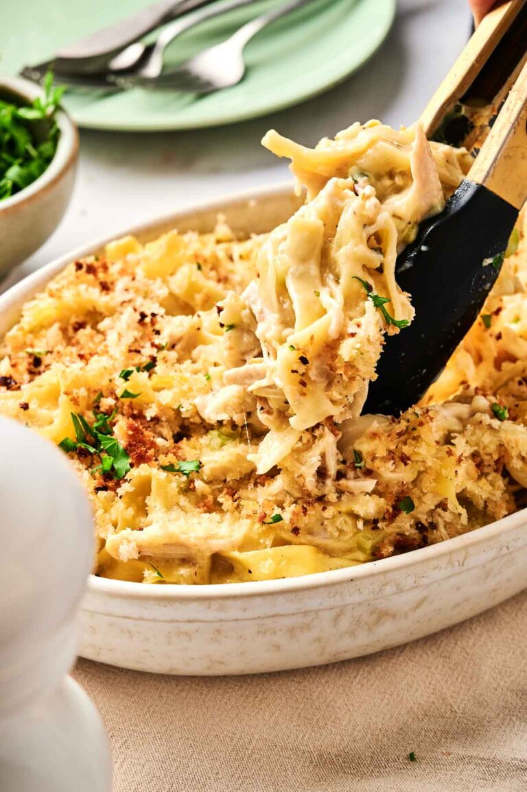 A serving of creamy chicken noodle casserole is being lifted from a white dish using tongs. The dish is garnished with chopped herbs and crumbs.