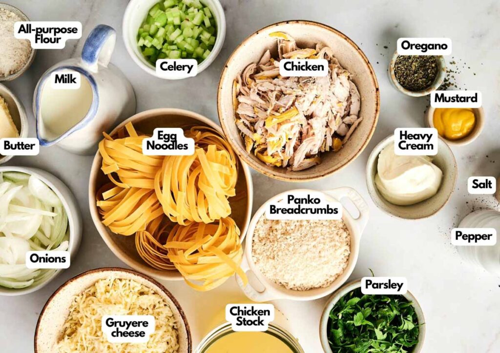 An assortment of labeled ingredients on a white surface, perfect for chicken noodle casserole, including shredded chicken, egg noodles, vegetables, cheese, milk, cream, spices, breadcrumbs, and chicken stock.