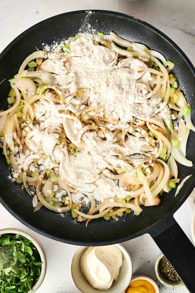 A pan with saut&eacute;ed onions and celery topped with flour, perfect for a savory chicken noodle casserole. Surrounding items include a bowl of herbs, a small bowl with a white substance, and a bowl of cracked egg.
