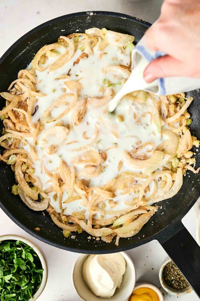 A hand pouring liquid over sliced onions in a skillet, with bowls of chicken noodle casserole ingredients nearby.