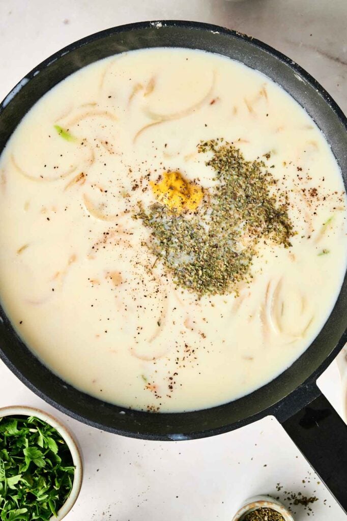 A pan filled with creamy chicken noodle casserole, topped with herbs and spices. Nearby is a small bowl of chopped greens.