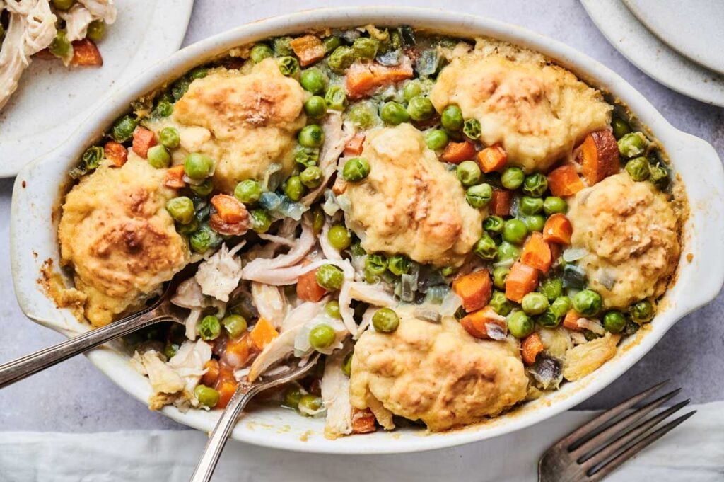 A baked chicken noodle casserole with vegetables, topped with biscuit dough, features peas and carrots visible in an oval dish and is served with two spoons.
