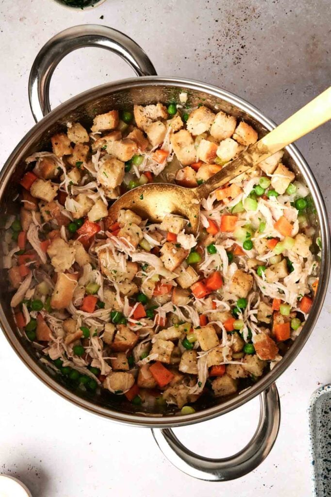 A pot filled with chicken and stuffing casserole, a hearty mix of cubed bread, shredded chicken, peas, carrots, and seasoning, with a gold spoon resting on top.