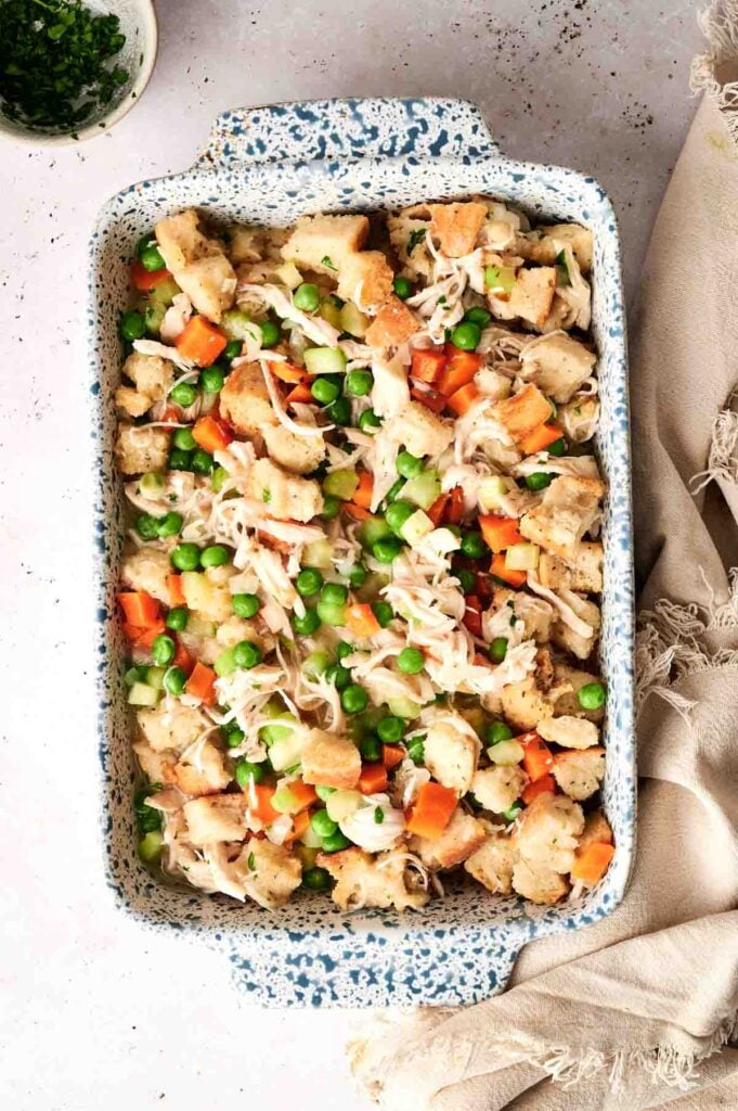 A chicken and stuffing casserole dish brimming with a mixture of bread cubes, shredded chicken, peas, carrots, and herbs rests on a light surface.