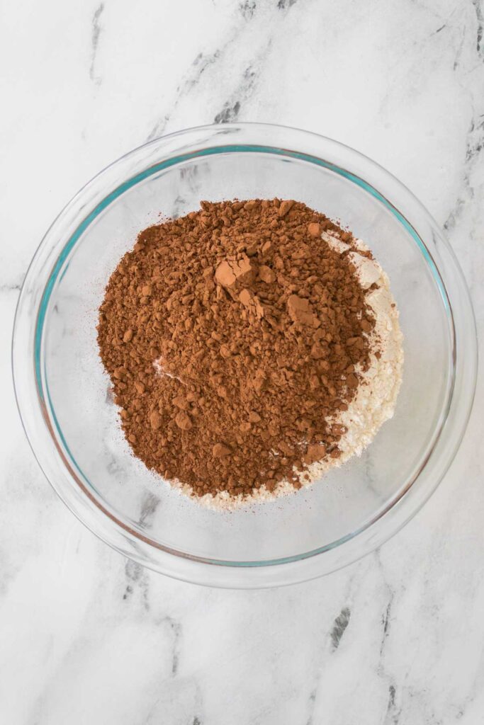 A glass bowl containing a rich cocoa powder and flour mix, perfect for baking chocolate cherry cookies, rests elegantly on a marble surface.