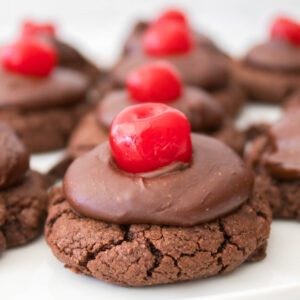 Chocolate cherry cookies with rich chocolate icing, each adorned with a vibrant red cherry, arranged on a pristine white plate.