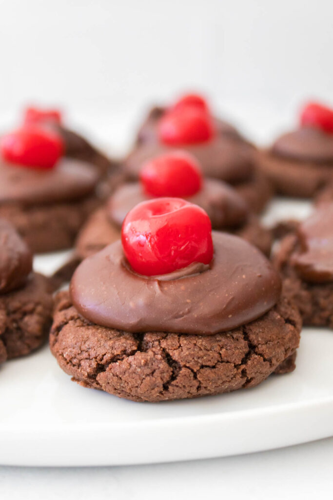 Chocolate cherry cookies with rich chocolate icing, each adorned with a vibrant red cherry, arranged on a pristine white plate.