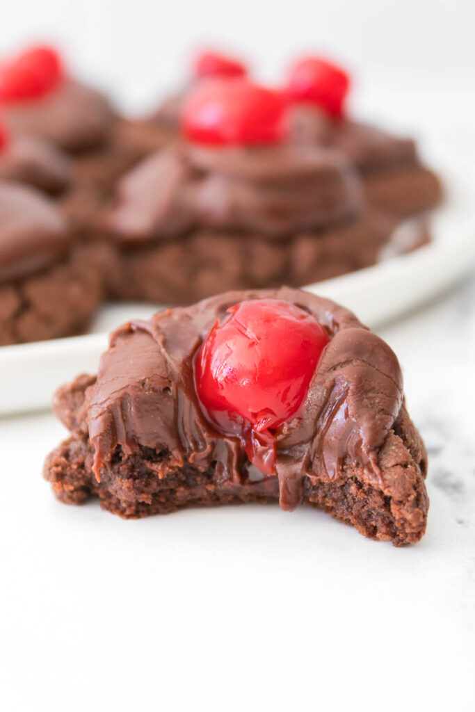 A delectable chocolate cherry cookie with a bite taken out sits enticingly in front of a plate brimming with more of these delightful treats.